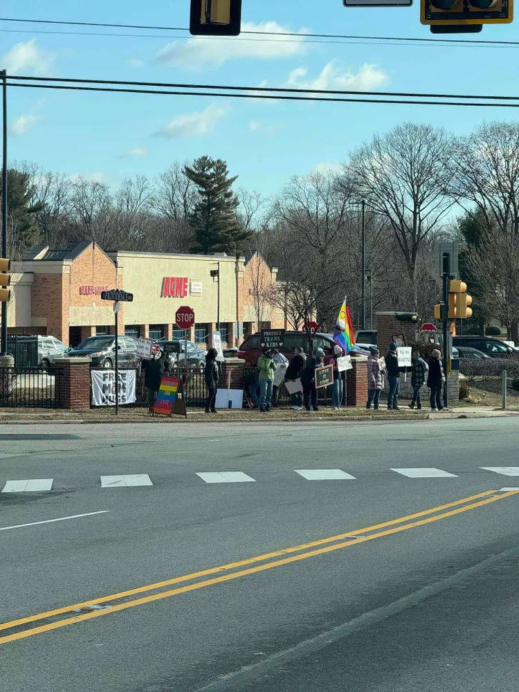 A photo of a group gathered on a corner. They carry signs such as “Protect Trans Kids” and “You Are Beautiful” and “Free Mom Hugs.”